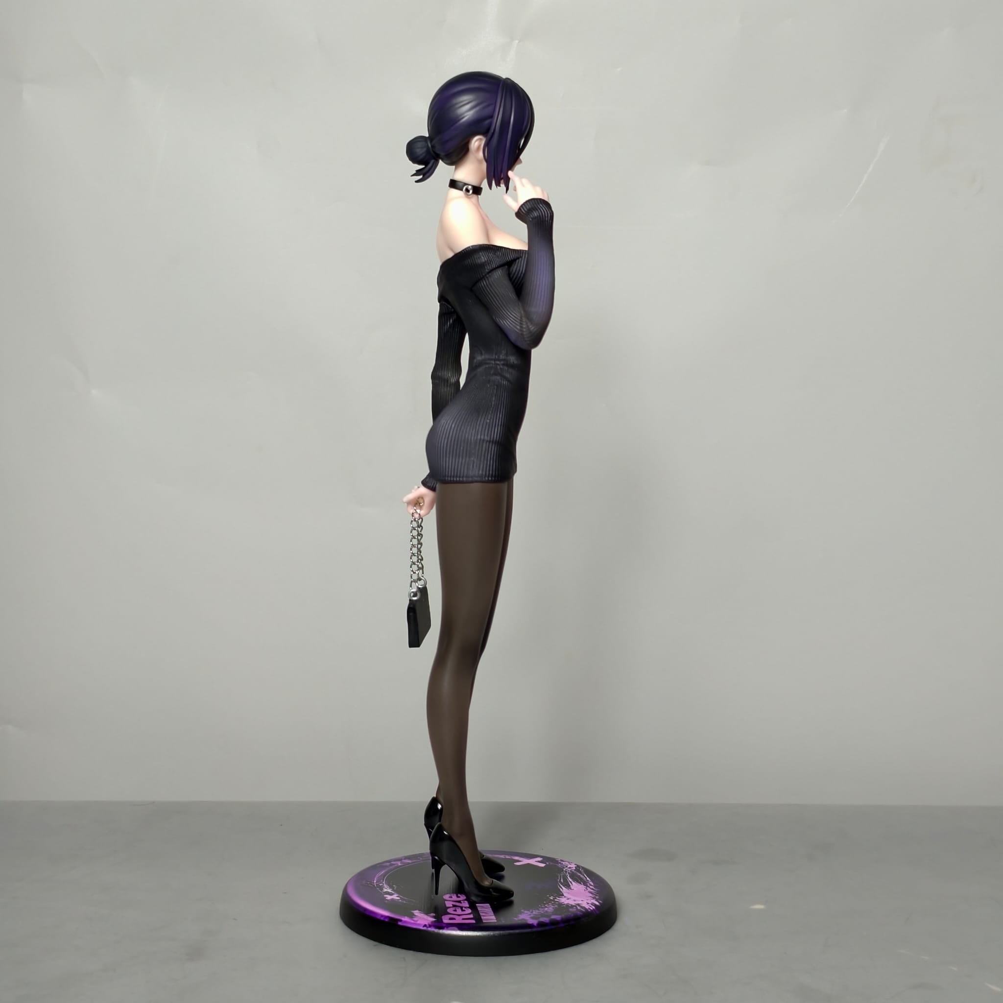 Vista laterale della figure di Reze in abito nero, con tacchi, borsetta e base espositiva viola e nera.